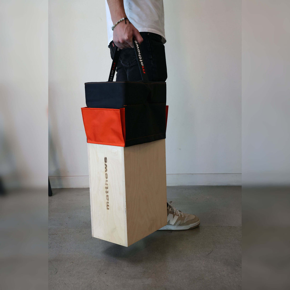 Person carrying a stacked Apple Box Topper™ in black and orange on a wooden Apple Box, showcasing its portable design.